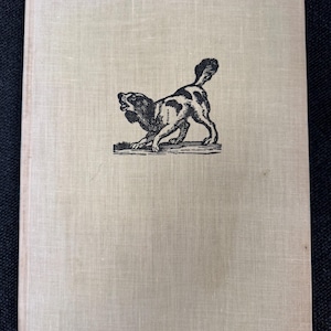 Könnte beinhalten: Ein Vintage-Buch mit hellbeigem Leinenumschlag. Eine Schwarz-Weiß-Illustration eines gefleckten Hundes befindet sich auf dem vorderen Einband. Der Hund befindet sich in einer spielerischen Haltung, mit erhobenem Schwanz und offenem Maul.