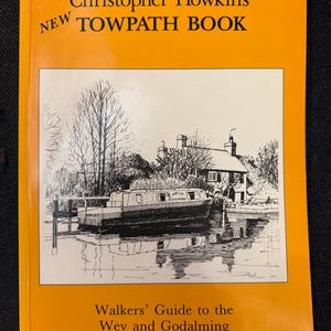 May include: A book titled "Christopher Howkins New Towpath Book" with an orange cover. The cover features a black and white illustration of a canal boat and a house. The text "Walkers' Guide to the Wey and Godalming Navigations" is also on the cover.