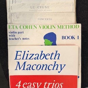 May include: A stack of sheet music and books, including "ETA Cohen Violin Method Book 1" and "Elizabeth Maconchy 4 easy trios." The covers feature text in English and German, with a blue, green, and red color scheme. The sheet music includes titles like "Meditation" and "Concerto."