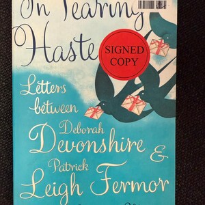 May include: A book cover with the title "In Tearing Haste" in elegant script over a turquoise background. The cover features the names Deborah Devonshire and Patrick Leigh Fermor, with a "Signed Copy" sticker.