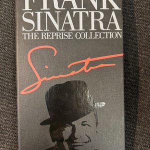 Puede incluir: Una caja de CD para "Frank Sinatra: The Reprise Collection" con fondo negro. El título está en letras blancas, con "Sinatra" en escritura roja. Se presenta una imagen en blanco y negro de Frank Sinatra con un sombrero fedora.