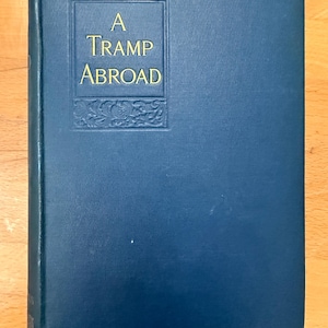 Puede incluir: Un libro de tapa dura titulado A Tramp Abroad con una cubierta texturizada de color azul oscuro. La portada presenta un panel rectangular central con bordes de motivos florales en relieve y el título A Tramp Abroad en letras doradas.