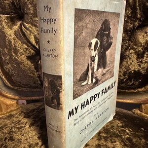 May include: A vintage book cover with a blue background and the title "My Happy Family" in black text. The book features a black and white photo of a chimpanzee and a dog. The author is Cherry Kearton. The book has 17 photographs.