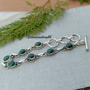 May include: A silver bracelet with a toggle clasp and green oval stones. The bracelet is on a grey surface.