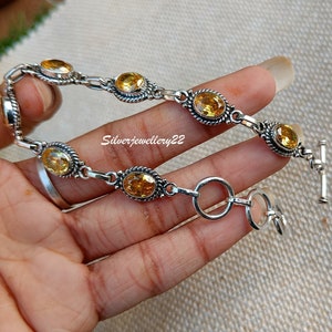 以下が含まれることがあります： チェーンリンクのデザインと楕円形の黄色の宝石が付いたシルバーブレスレット。ブレスレットはトグル留め具です。