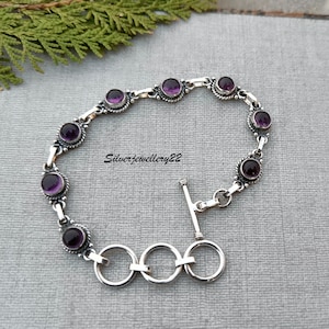 Pode incluir: Uma pulseira de prata com fecho de correr e oito acentos de pedras preciosas roxas. A pulseira é feita de prata esterlina e apresenta um design de corrente delicado.