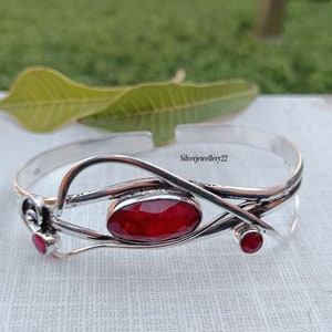 Könnte beinhalten: Ein silbernes Armreif-Armband mit einem roten Edelstein und zwei kleineren roten Edelsteinen. Das Armband hat ein zartes, filigranes Design.