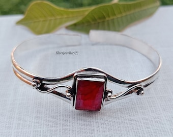 Puede incluir: Un brazalete de plata con una piedra preciosa roja engastada en un marco de plata. El brazalete tiene un diseño delicado y en espiral.