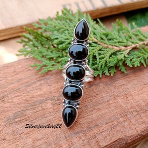 Peut inclure: Une bague en argent avec cinq pierres d'onyx noir disposées en rangée verticale. La bague est posée sur une surface en bois.