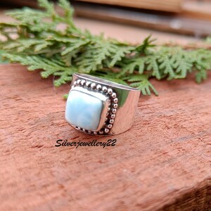 May include: A silver ring with a square blue gemstone set in a beaded bezel. The ring is on a wooden surface.