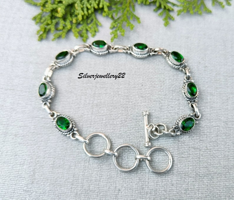May include: A silver bracelet with a toggle clasp and green oval-shaped gemstones. The bracelet is on a gray background.