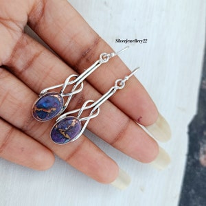 Puede incluir: Un par de pendientes colgantes de plata con piedras ovaladas de color morado y cobre. Los pendientes tienen un diseño de nudo y miden aproximadamente 5 cm de largo.
