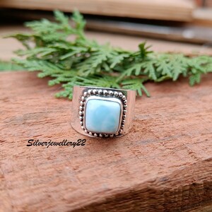 May include: A silver ring with a square blue gemstone set in a beaded bezel. The ring is on a wooden surface.