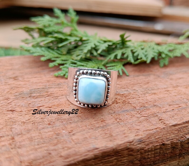 May include: A silver ring with a square blue gemstone set in a beaded bezel. The ring is on a wooden surface.