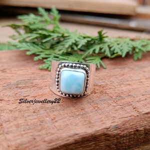 May include: A silver ring with a square blue gemstone set in a beaded bezel. The ring is on a wooden surface.