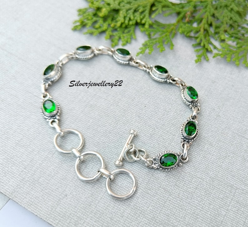 May include: A silver bracelet with a toggle clasp and green oval-shaped gemstones. The bracelet is on a gray background.