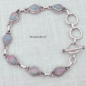 Puede incluir: Un brazalete de plata con cierre de palanca y seis piedras ovaladas iridiscentes. El brazalete está sobre un fondo de tela blanca.