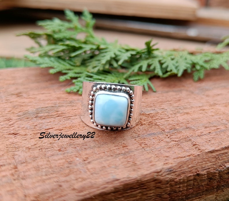 May include: A silver ring with a square blue gemstone set in a beaded bezel. The ring is on a wooden surface.