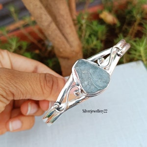 May include: A silver bangle bracelet featuring a green gemstone set in the centre. The bracelet has a delicate, swirling design.