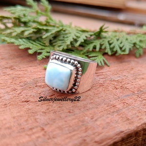 May include: A silver ring with a square blue gemstone set in a beaded bezel. The ring is on a wooden surface.