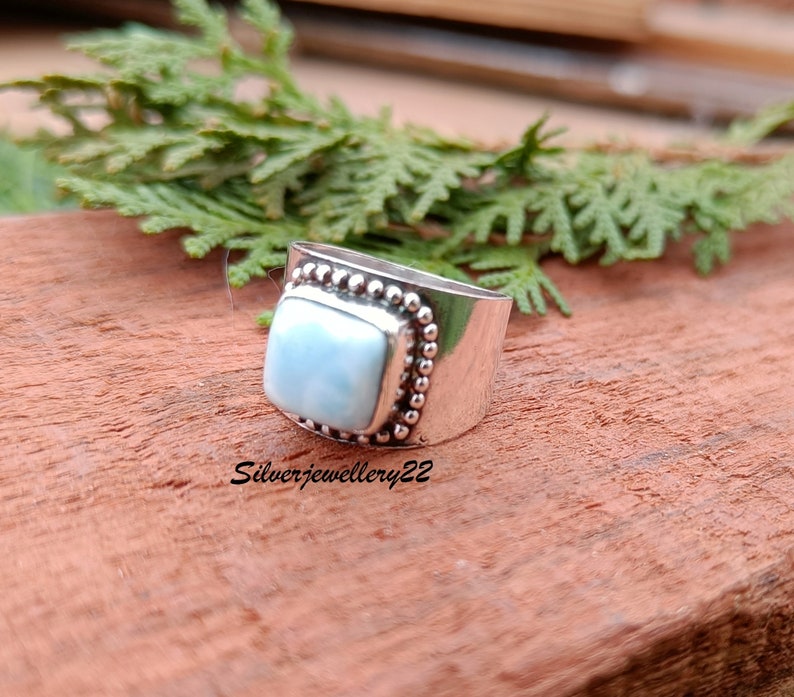 May include: A silver ring with a square blue gemstone set in a beaded bezel. The ring is on a wooden surface.