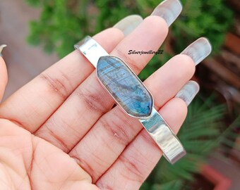 May include: A silver bangle bracelet featuring a large, iridescent labradorite gemstone set in the centre. The gemstone is a deep blue with flashes of green and gold.