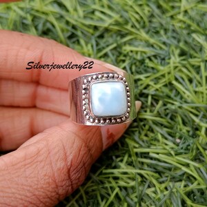 May include: A silver ring featuring a square, light blue gemstone set within a beaded bezel. The ring is displayed on a finger against a green backdrop.
