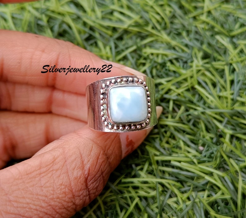 May include: A silver ring featuring a square, light blue gemstone set within a beaded bezel. The ring is displayed on a finger against a green backdrop.