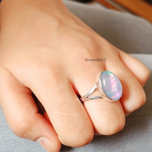 May include: A silver ring with an oval, iridescent opal gemstone. The ring's band splits into two delicate prongs that cradle the stone. The ring is worn on a finger, showcasing its design and the play of colors within the opal.