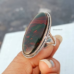 Puede incluir: Un anillo de plata con una gran piedra ovalada de color verde, rojo y marrón. El anillo tiene un diseño de cuerda alrededor del borde del engaste.