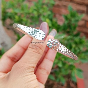 May include: A silver cuff bracelet with a hammered texture. The open design features a slightly flattened, rounded shape. The bracelet is held against a background of green foliage and a brick wall.