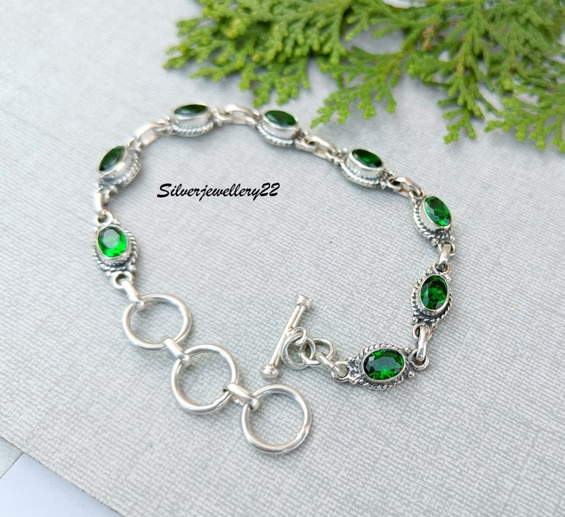 May include: A silver bracelet with a toggle clasp and green oval-shaped gemstones. The bracelet is on a gray background.