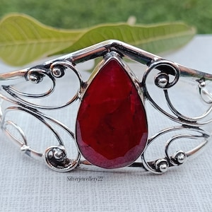 Può includere: Un braccialetto d'argento con un grande rubino a forma di goccia incastonato al centro. Il braccialetto ha un design delicato e ornato con volute e curve.