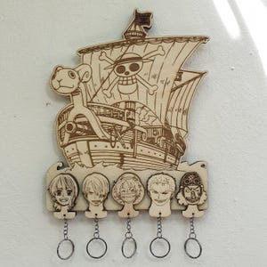 Op de afbeelding: Houten sleutelhouder met een scheepsontwerp met een doodshoofd en gekruiste botten, en vijf gezichten van personages. Elk gezicht van een personage heeft een metalen sleutelring. De sleutelhouder is gemaakt van lichtbruin hout.