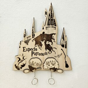 Puede incluir: Porta llaves de madera con una silueta del castillo de Hogwarts, las palabras "Expecto Patronum" y dos personajes de Harry Potter.