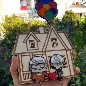 Può includere: Un ritaglio di legno di una casa con una coppia in piedi davanti. La casa è marrone e ha un cuore rosso sopra la coppia. La coppia si tiene per mano e ha una catena attaccata ai polsi. La casa è attaccata a un mazzo di palloncini colorati.