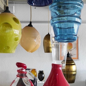 Op de afbeelding: Een assortiment van kleurrijke glazen hanglampen en vazen. De lampen hebben verschillende vormen en maten, waaronder een blauwe gelaagde glazen kap, een gele glazen kap met gaten en een crèmekleurige glazen kap. De vazen zijn van rood en helder glas met gouden accenten.