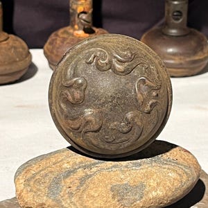 May include: A vintage, ornate, dark brown metal door knob with a decorative floral design. The knob is sitting on a rough, gray and brown stone.