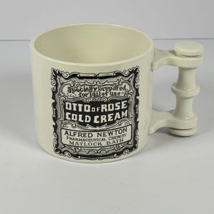 May include: Cream-colored ceramic mug with a unique handle design. The mug features a black and white label with the text "OTTO OF ROSE COLD CREAM" and "ALFRED NEWTON PHARMACEUTICAL CHEMIST MATLOCK BATH".