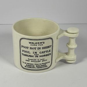 May include: Cream-colored ceramic mug with a unique handle design. The mug features black text within a decorative border, advertising a cure for foot rot in sheep, foul in cattle, and thrush in horses. The text also mentions the proprietor, Aaron Walker, and the location.