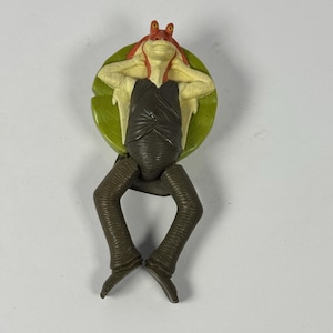 May include: A plastic action figure of a character from a science fiction film. The figure has a red head, yellow skin, and a black outfit. It is posed on a green lily pad and has brown legs.