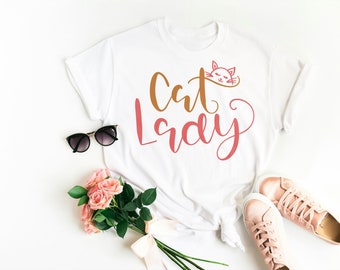 Peut inclure: T-shirt blanc avec le texte "Cat Lady" en lettres brunes et roses. Un petit graphique de visage de chat est au-dessus du texte. Le t-shirt est posé à plat sur une surface blanche avec des lunettes de soleil, des roses roses et des baskets roses.
