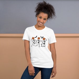 May include: White t-shirt featuring a Halloween design. The graphic includes three dancing skeletons with pumpkin heads and black bats. The skeletons are black and white, and the pumpkins are orange. The shirt is a crew neck.