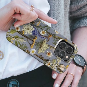 May include: A gray phone case featuring a floral design with pressed flowers in white, yellow, and blue. The case has a glossy finish and a camera cutout. The phone case is held by a person.