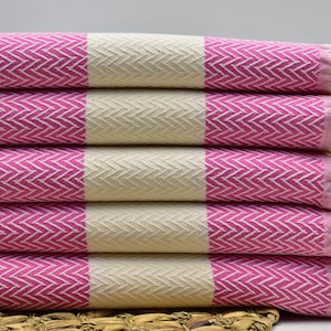 Puede incluir: Una pila de toallas rayadas de color rosa y blanco con flecos en los bordes. Las toallas están dobladas por la mitad y apiladas una encima de la otra.