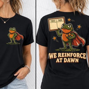 May include: A black t-shirt featuring a graphic of a green frog wearing a red cape. The front shows the frog standing, while the back displays the frog holding a sign that reads FIRST THEN and the text WE REINFORCE AT DAWN with star icons.