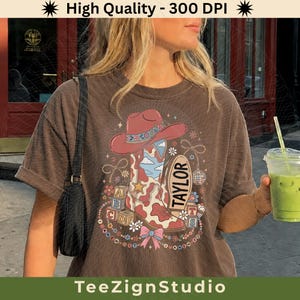 May include: A brown short-sleeve t-shirt featuring a graphic print of a pink cowboy hat, a cow-print cowboy boot, alphabet blocks, a disco ball, and a bow. The text TAYLOR is printed on an oval shape. The design includes stars and flowers.