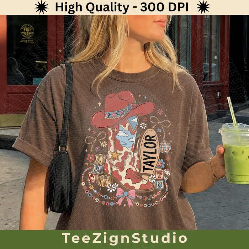 May include: A brown short-sleeve t-shirt featuring a graphic print of a pink cowboy hat, a cow-print cowboy boot, alphabet blocks, a disco ball, and a bow. The text TAYLOR is printed on an oval shape. The design includes stars and flowers.