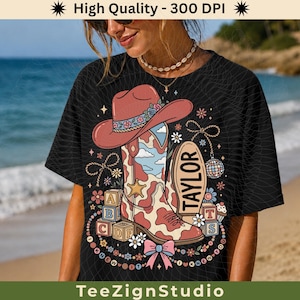 May include: A black t-shirt featuring a graphic print of a pink cowboy hat, a cow-print cowboy boot, alphabet blocks, a disco ball, and flowers. The text TAYLOR is printed on a vertical oval shape next to the boot, surrounded by small stars.