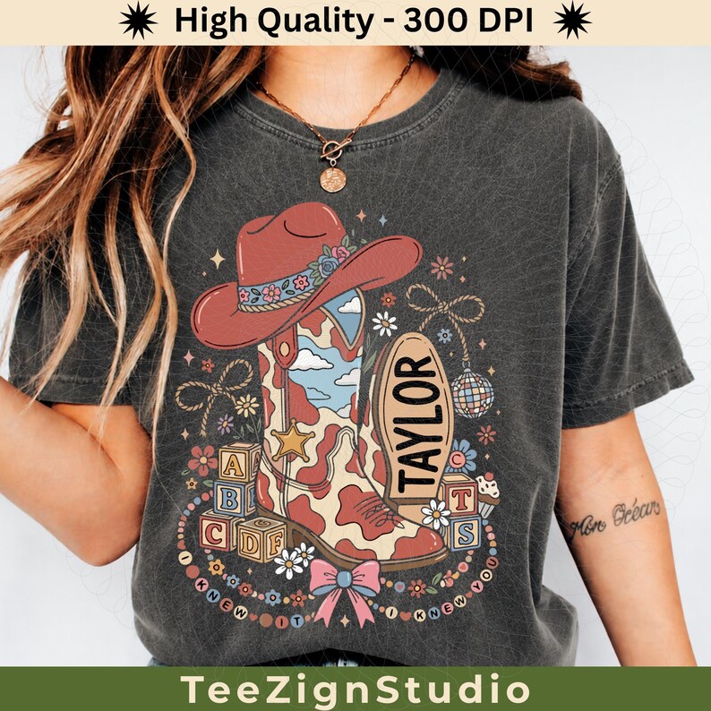 May include: A charcoal gray t-shirt featuring a graphic of a red cowboy hat and a cow-print boot with a blue sky pattern. The design includes alphabet blocks, a disco ball, flowers, a bow, and the text "TAYLOR" and "I KNEW IT I KNEW YOU".